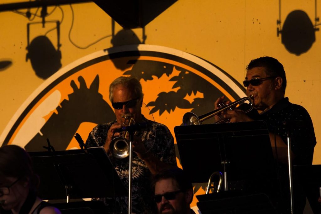   Members or the Milwaukee Jazz Orchestra play trumpets as the sun casts shadows on the stage backdr